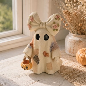 May include: A cream-coloured ghost figurine featuring a large bow on its head decorated with pink floral patterns. The ghost has black circular eyes, a patchwork-style body with various coloured fabric textures, and holds a small orange pumpkin bucket.