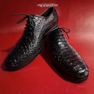 Pode incluir: Um par de sapatos oxford pretos com atacadores, apresentando um padrão texturizado semelhante a escamas no exterior. Os sapatos têm biqueira arredondada, atacadores pretos finos e detalhes brogue subtis nos painéis laterais. O texto ACALAPATIVN é visível.