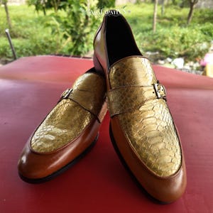 Pu&ograve; includere: Un paio di scarpe con fibbia caratterizzate da una base in pelle marrone chiaro liscia e una tomaia con texture in pelle di serpente color oro metallizzato. Ogni scarpa presenta una fibbia in metallo argentato su un cinturino e una suola scura.