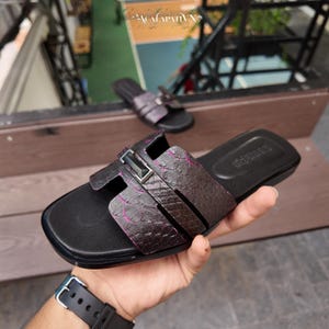 May include: A pair of dark purple and black slide sandals featuring a textured snakeskin pattern on the wide strap. The strap is accented with a central rectangular metallic hardware piece. The footbed is black with a smooth finish.