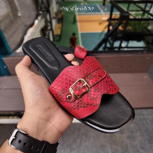 May include: A slide sandal featuring a black footbed and a wide, red snakeskin-textured strap across the top. The strap is accented with a gold-toned metal buckle and a small metal stud. The brand name ACALAPATI VN is embossed on the black insole.