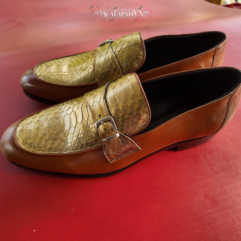 Pu&ograve; includere: Un paio di mocassini in pelle marrone con tomaia in texture di pelle di serpente color oro metallizzato e cinturino decorativo con fibbia in metallo color argento. Le scarpe presentano una finitura in pelle marrone liscia e fodera nera.