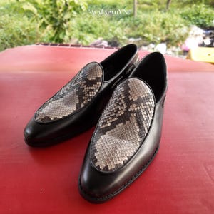Pode incluir: Um par de mocassins sem atacadores com estrutura em pele preta e gáspea texturizada com padrão de pele de cobra cinzento e preto. Os sapatos têm biqueira arredondada e silhueta de perfil baixo, mostrando um contraste entre a pele lisa e o padrão detalhado de escamas na parte superior.