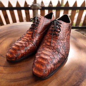 Peut inclure: Une paire de chaussures richelieu marron avec un motif texturé en peau de serpent, des lacets noirs et un bout droit avec des détails brogues subtils. Les chaussures ont une finition polie et une semelle sombre, avec un aspect écailleux.