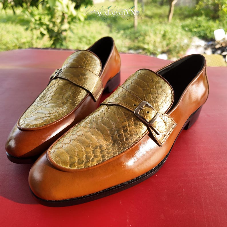 Pu&ograve; includere: Un paio di mocassini con base in pelle liscia color cuoio e tomaia testurizzata con motivo a pelle di serpente color oro. Ogni scarpa presenta un cinturino decorativo sul collo del piede fissato da una piccola fibbia metallica e un tacco basso.