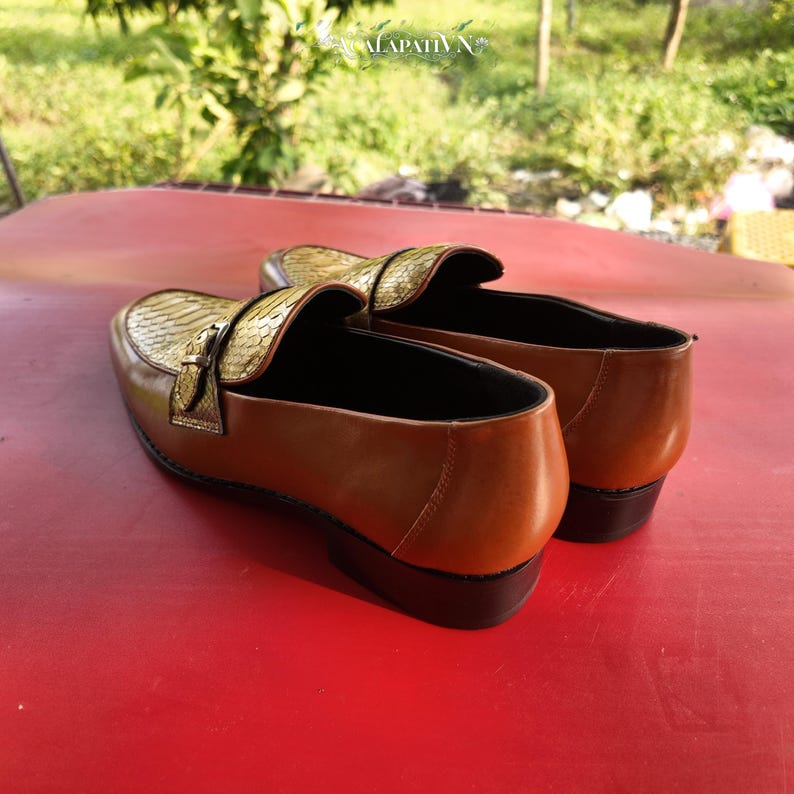 Pu&ograve; includere: Un paio di mocassini slip-on con tallone e pannello laterale in pelle liscia color cuoio, a contrasto con una tomaia testurizzata con motivo a pelle di serpente dorata. Ogni scarpa presenta un cinturino decorativo con fibbia in metallo dorato.