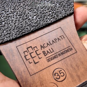 May include: The brown sole of a shoe features a black textured tread and a stamped logo reading ACALAPATI BALI, DESIGNED IN INDONESIA, with the number 35 enclosed in a circle. The wood-toned surface displays a clear brand mark and size indicator.