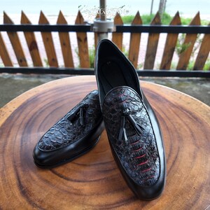 Pode incluir: Um par de mocassins pretos com um padrão texturizado semelhante a escamas no peito do pé com um subtil tom avermelhado. Cada sapato é adornado com um detalhe de borla preta na frente. O design inclui uma borda de couro preto liso.