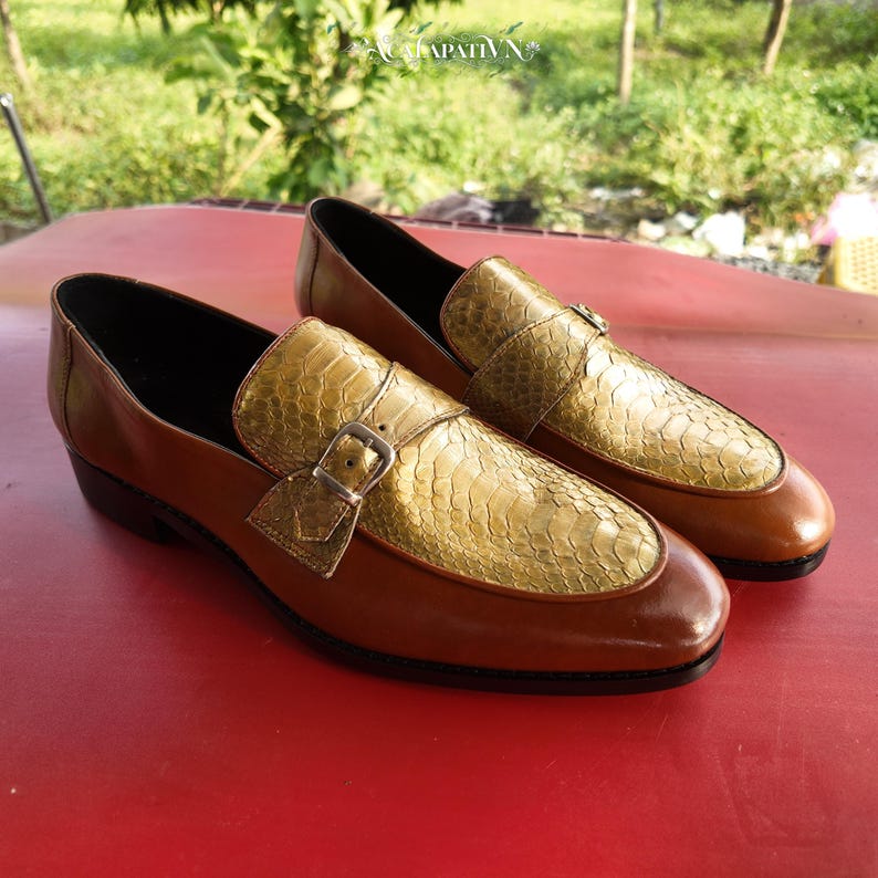 Pu&ograve; includere: Un paio di mocassini in pelle marrone con un pannello frontale dalla texture in pelle di serpente color oro metallizzato. Ogni scarpa presenta un cinturino decorativo sul collo del piede fissato con una piccola fibbia in metallo argentato. La fodera &egrave; nera.