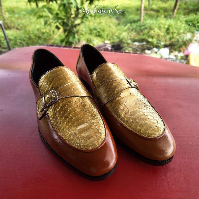 Pu&ograve; includere: Un paio di mocassini in pelle marrone con tomaia dorata dalla texture effetto pelle di serpente e un cinturino decorativo con fibbia in metallo color argento sulla parte superiore. Le scarpe presentano una finitura liscia e punta tonda.