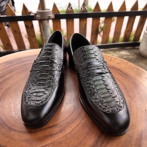Pode incluir: Um par de mocassins pretos com um peito do pé texturizado com padrão de pele de cobra e detalhes verdes subtis visíveis entre as escamas. Os sapatos têm uma biqueira e calcanhar em couro liso, uma tira clássica e um design de bico redondo.