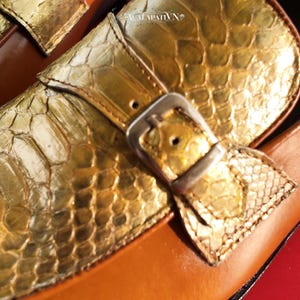 Pu&ograve; includere: Un paio di mocassini con tomaia in pelle di serpente dorata metallizzata su base in pelle marrone. Un cinturino in pelle di serpente dorata attraversa la parte superiore, fissato da una fibbia in metallo color argento. Testo ACALAPATI VN visibile.