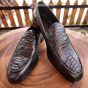 Pode incluir: Um par de mocassins pretos sem atacadores com um padrão texturizado semelhante a escamas no peito do pé e detalhes vermelhos subtis entre as escamas. Os sapatos têm um acabamento em pele lisa nas laterais e um salto baixo.