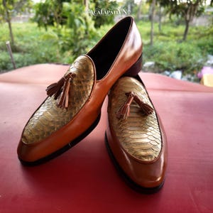 Pode incluir: Um par de mocassins de couro castanho com um peito do pé texturizado com padrão de pele de cobra dourado metálico e uma borla decorativa em cada sapato. Os sapatos têm um acabamento em couro liso, biqueira arredondada e sola escura, com o texto ACALAPATYN visível.