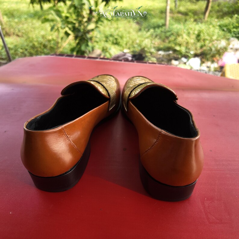 Pu&ograve; includere: Un paio di mocassini slip-on con talloni e quartieri in pelle liscia color cuoio e tomaie dorate testurizzate. Le scarpe presentano interni neri e tacchi a blocco marrone scuro. Il testo ACALAPATI VN &egrave; visibile nella parte superiore centrale.
