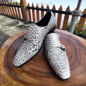 May include: A pair of double monk strap shoes featuring a textured, grey and white snakeskin pattern. The shoes have a pointed toe, a black interior lining, and a black sole. Each shoe is secured with two silver-toned metal buckles on the side.