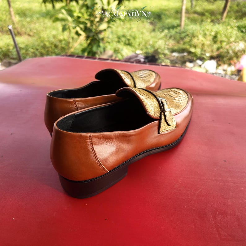 Pu&ograve; includere: Un paio di mocassini in pelle marrone con tomaia dorata dalla texture effetto pelle di serpente e un cinturino decorativo con fibbia metallica sulla parte superiore. Le scarpe hanno un tacco basso nero e una finitura liscia, con il marchio ACALAPATI VN.