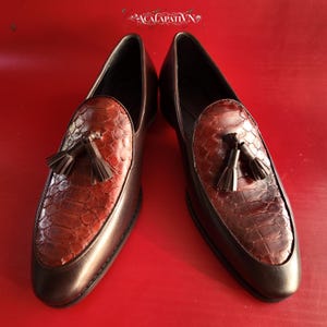 Pode incluir: Um par de mocassins castanho-escuros com um peito do pé texturizado com padrão de pele de cobra castanho-avermelhado e um detalhe de borla decorativa no topo. Os sapatos têm um acabamento em pele lisa e um design de biqueira pontiaguda.