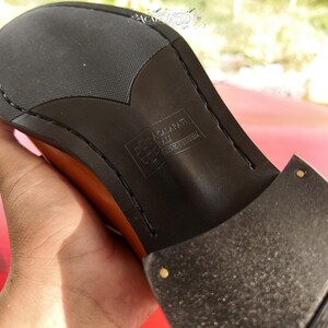 Pu&ograve; includere: La suola nera di una scarpa color cuoio con un motivo a battistrada testurizzato e un tacco a strati. Al centro della suola &egrave; impresso il testo ACALAPATI BALI DESIGNED IN INDONESIA, accompagnato da un piccolo logo quadrato.