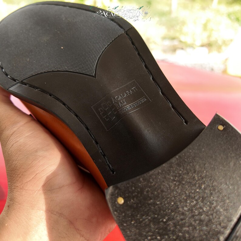 Pu&ograve; includere: La suola nera di una scarpa color cuoio con un motivo a battistrada testurizzato e un tacco a strati. Al centro della suola &egrave; impresso il testo ACALAPATI BALI DESIGNED IN INDONESIA, accompagnato da un piccolo logo quadrato.