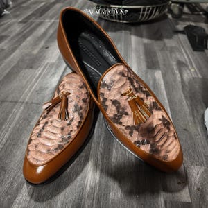 May include: A pair of tan leather loafers featuring a snakeskin-textured vamp panel, a gold-toned tassel ornament on each shoe, and a black interior lining. The brand name ACALAPATI VN is visible on the inner sole and at the top of the image.
