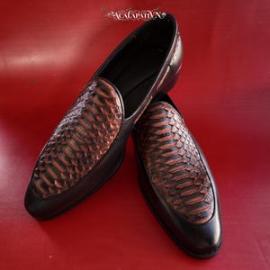 Peut inclure: Une paire de mocassins à enfiler avec une structure en cuir noir et un empeigne texturé à motif peau de serpent marron. Les chaussures présentent un bout pointu et une doublure intérieure noire lisse. La marque ACALAPATI VN est visible.