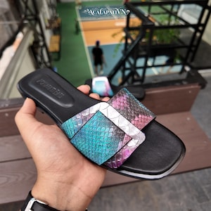 May include: A black slide sandal featuring a wide strap with a snakeskin texture in a gradient of teal, purple, and pink. The strap includes a large, silver-toned rectangular buckle detail. The footbed is black with a debossed logo on the heel area.