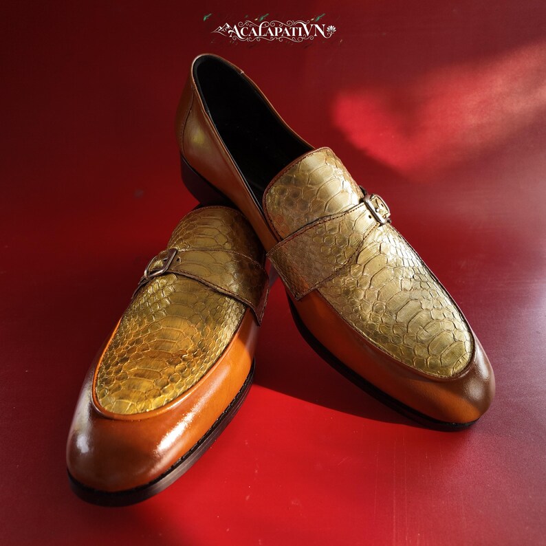 Pu&ograve; includere: Un paio di mocassini in pelle color cuoio con una tomaia testurizzata a motivo pelle di serpente dorata e un cinturino singolo con una piccola fibbia in metallo. Le scarpe presentano una finitura in pelle liscia, fodera scura e il testo ACALAPATIVN.