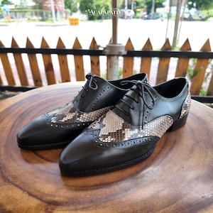 Pode incluir: Um par de sapatos oxford de couro preto com design wingtip, detalhes brogue e um painel com padrão de pele de cobra no peito do pé e no calcanhar. Os sapatos têm atacadores pretos, biqueira pontiaguda e um salto baixo, com visual bicolor.