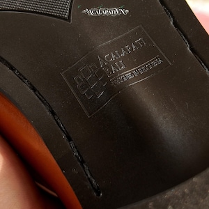Pu&ograve; includere: La suola nera di una scarpa con un motivo del battistrada testurizzato e un logo rettangolare in rilievo. Il logo mostra il testo ACALAPATI BALI e DESIGNED IN INDONESIA. Un piccolo logo decorativo con il testo ACALAPATI VN &egrave; visibile in alto.