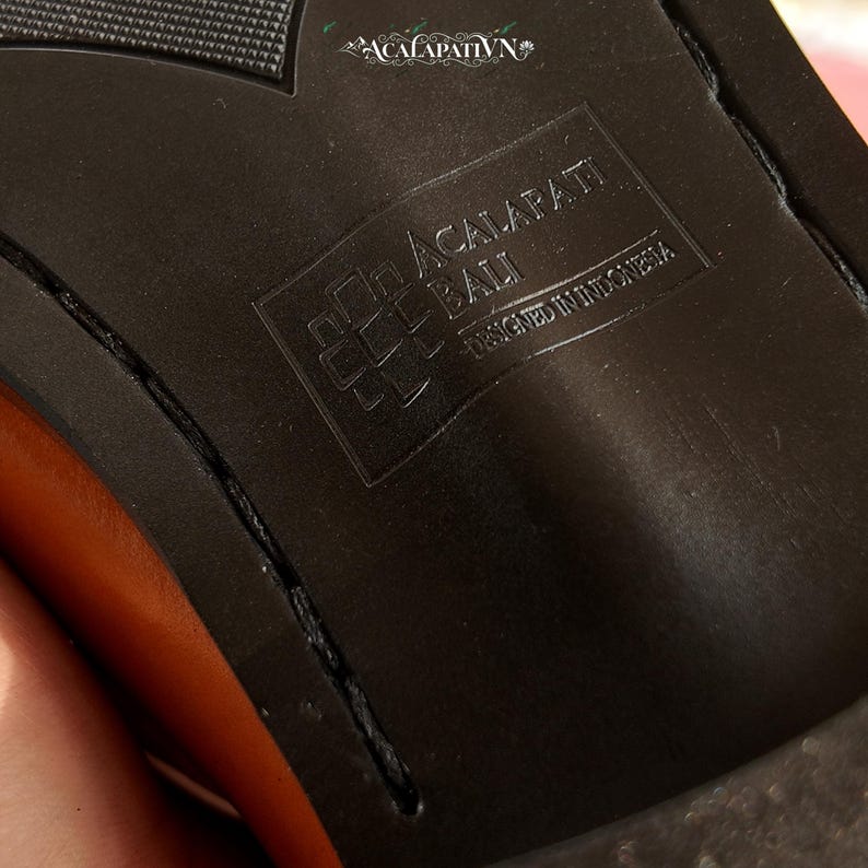 Pu&ograve; includere: La suola nera di una scarpa con un motivo del battistrada testurizzato e un logo rettangolare in rilievo. Il logo mostra il testo ACALAPATI BALI e DESIGNED IN INDONESIA. Un piccolo logo decorativo con il testo ACALAPATI VN &egrave; visibile in alto.