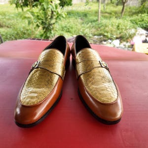 Pu&ograve; includere: Un paio di mocassini con base in pelle marrone liscia e tomaia con texture in pelle di serpente color oro metallizzato. Ogni scarpa presenta un cinturino decorativo sulla parte superiore con una piccola fibbia in metallo argentato e suola nera.