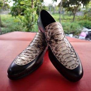 May include: A pair of black leather loafers featuring a textured, light gray and black snakeskin-patterned vamp panel. The shoes have a rounded toe, a low heel, and decorative brogue-style perforated detailing along the edge of the snakeskin inlay.