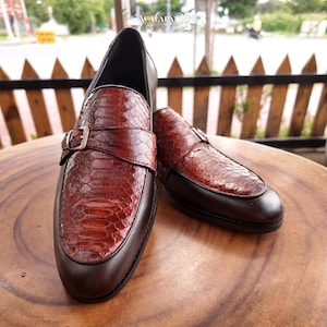 May include: A pair of brown loafers featuring a textured, reddish-brown snakeskin-patterned vamp and a silver-toned metal buckle strap across the top. The shoes have a smooth, dark brown leather toe cap and heel section with a rounded toe profile.