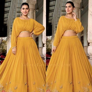 May include: A mustard yellow two-piece set consisting of a cropped long-sleeve blouse with a round neckline and a floor-length pleated skirt. The skirt features intricate floral embroidery in shades of red, green, and white along the hemline.