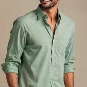May include: A sage green long-sleeved button-down shirt featuring a classic collar, a single chest pocket, and light-colored buttons. The shirt is crafted from a smooth fabric and is worn with the sleeves rolled up to the mid-forearm.