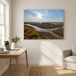 May include: A rectangular wall art print depicting a winding tidal creek through golden-brown marshland under a bright sun and blue sky with wispy clouds. The landscape scene is mounted on a plain wall above a wooden desk and a beige armchair.