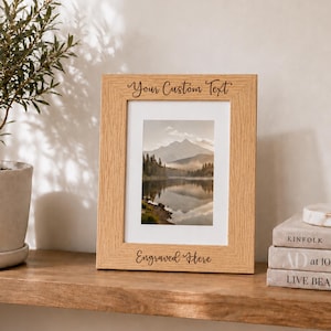 May include: A rectangular light brown wooden picture frame with a visible wood grain texture. The frame features the text Your Custom Text engraved at the top and Engraved Here at the bottom, surrounding a vertical photograph of a mountain lake.