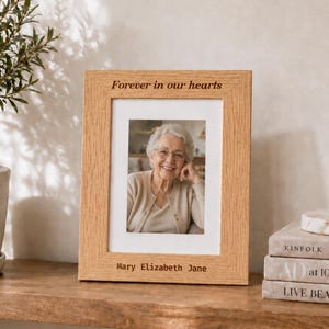 May include: A rectangular light brown wooden picture frame with a visible grain texture. The top of the frame is engraved with the text "Forever in our hearts" and the bottom with "Mary Elizabeth Jane". It holds a portrait of an older person.