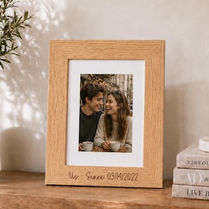 May include: A light oak wood picture frame featuring a white mat and a photograph of a couple holding mugs. The bottom of the frame is engraved with the text "Us · Since 03/14/2022" in a dark, cursive script font, centered below the photo.