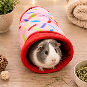 May include: A soft, cylindrical fleece tunnel for small animals featuring a light pink exterior with a colorful sprinkle pattern and a solid red interior lining. A grey and white guinea pig is positioned inside the open end of the tunnel.