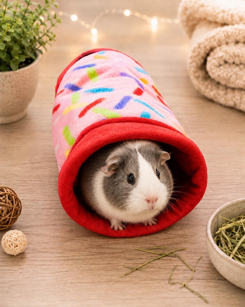 May include: A soft, cylindrical fleece tunnel for small animals featuring a light pink exterior with a colorful sprinkle pattern and a solid red interior lining. A grey and white guinea pig is positioned inside the open end of the tunnel.