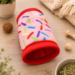 May include: A cylindrical tunnel for small animals made of soft pink fleece fabric featuring a colorful sprinkle pattern in blue, green, yellow, and red. The tunnel has solid red fleece trim at both ends and a solid red interior lining.