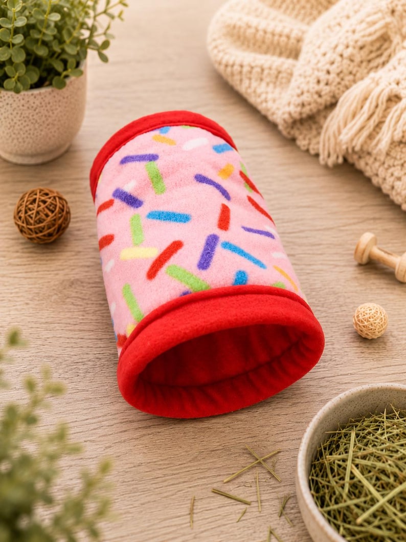 May include: A cylindrical tunnel for small animals made of soft pink fleece fabric featuring a colorful sprinkle pattern in blue, green, yellow, and red. The tunnel has solid red fleece trim at both ends and a solid red interior lining.