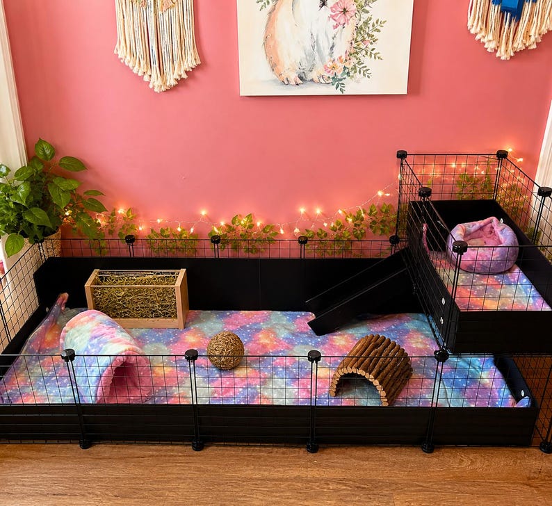 May include: A black wire pet cage with a multi-level design, featuring a ramp, a loft, and a colorful galaxy-patterned floor liner. The enclosure includes a wooden hay rack, a woven ball, a wooden tunnel, and a plush tie-dye bed.