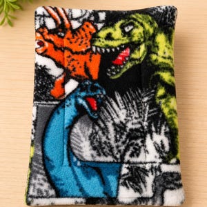 K&ouml;nnte beinhalten: Ein rechteckiger Stoffbeutel mit einem lebendigen Dinosaurier-Druck. Das Design zeigt einen gr&uuml;nen T-Rex, einen orangefarbenen Triceratops und einen blauen Langhals-Dinosaurier vor einem schwarz-wei&szlig;en Hintergrund mit detailliertem pr&auml;historischem Blattwerk.