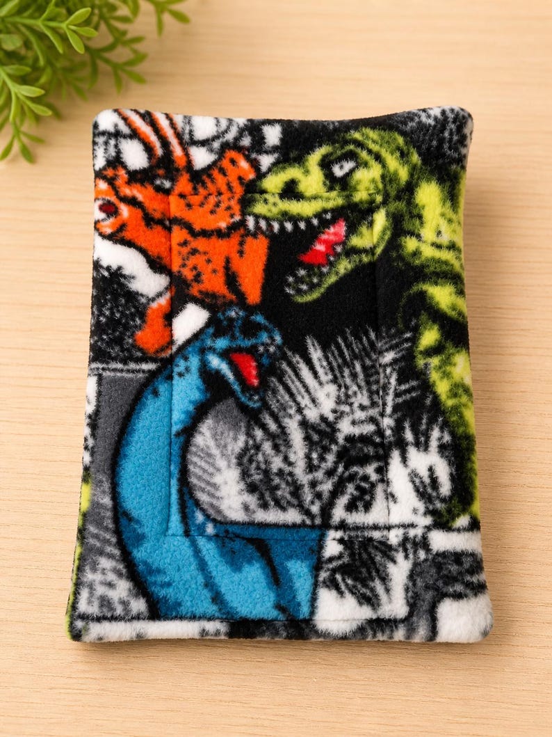 K&ouml;nnte beinhalten: Ein rechteckiger Stoffbeutel mit einem lebendigen Dinosaurier-Druck. Das Design zeigt einen gr&uuml;nen T-Rex, einen orangefarbenen Triceratops und einen blauen Langhals-Dinosaurier vor einem schwarz-wei&szlig;en Hintergrund mit detailliertem pr&auml;historischem Blattwerk.