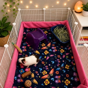 May include: A white wire pet enclosure with pink fabric liners containing a guinea pig on a dark blue fleece blanket with a colorful butterfly and flower pattern. The enclosure includes a purple plastic hide, a blue hay feeder, and toys.