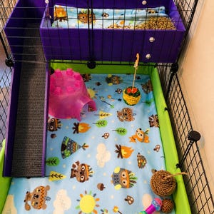 May include: A multi-level pet cage featuring a purple upper loft with a ramp, a light blue fleece liner with a woodland animal print, a pink plastic igloo hideout, a hanging yellow pom-pom toy, and a brown woven ball toy on the floor.
