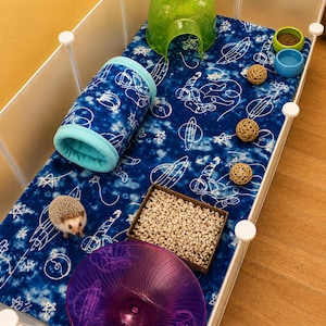 May include: A hedgehog enclosure featuring a blue fleece liner with white space-themed prints, a green plastic igloo, a blue fabric tunnel, a purple exercise wheel, a square wooden box filled with bedding, two small bowls, and three rattan balls.
